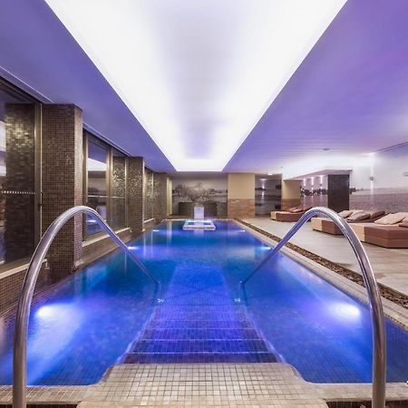 Ap Lago Montargil - Conference & Spa Zewnętrze zdjęcie The photo shows a modern indoor swimming pool area. The pool features a sleek design with steps leading into the water and a gentle curve at the sides. The water is illuminated in a blue hue, enhancing the relaxing atmosphere. Surrounding the pool ar