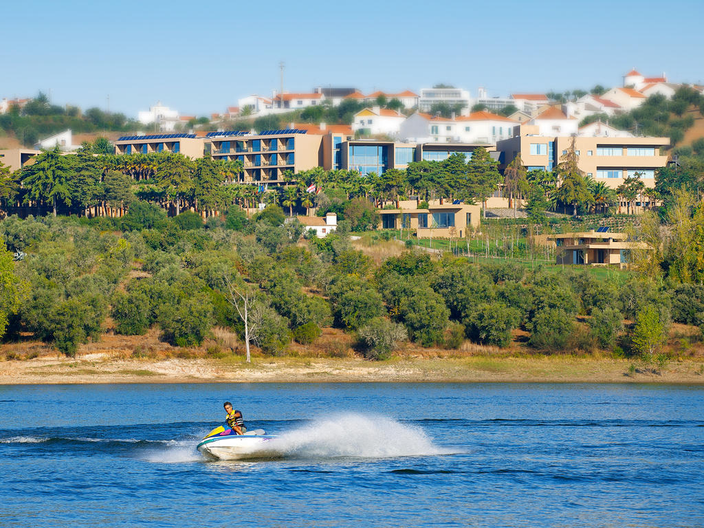 Ap Lago Montargil - Conference & Spa Zewnętrze zdjęcie The photo depicts a person riding a jet ski on a body of water. In the background, there are modern buildings and lush greenery, suggesting a scenic landscape, likely near a resort or upscale residential area. The clear blue sky and calm water enhanc