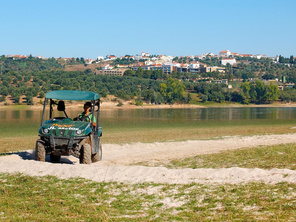 Ap Lago Montargil - Conference & Spa Zewnętrze zdjęcie The photo shows a small green all-terrain vehicle driving along a sandy path beside a body of water. In the background, there's a landscape with trees and rolling hills, along with a town or village visible on the hillside. The sky is clear, indicati