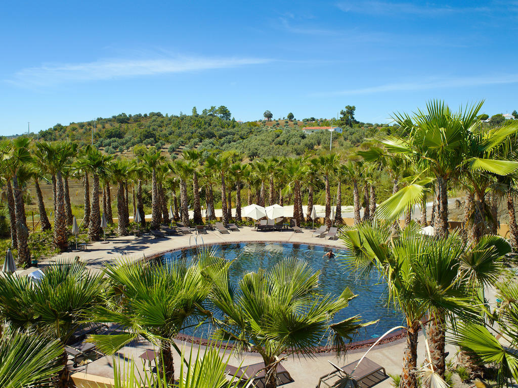 Ap Lago Montargil - Conference & Spa Zewnętrze zdjęcie The photo depicts a beautiful outdoor area featuring a circular swimming pool surrounded by palm trees. The pool area has several lounge chairs arranged around it. In the background, there is a lush green hillside, and the sky is clear with a bright 