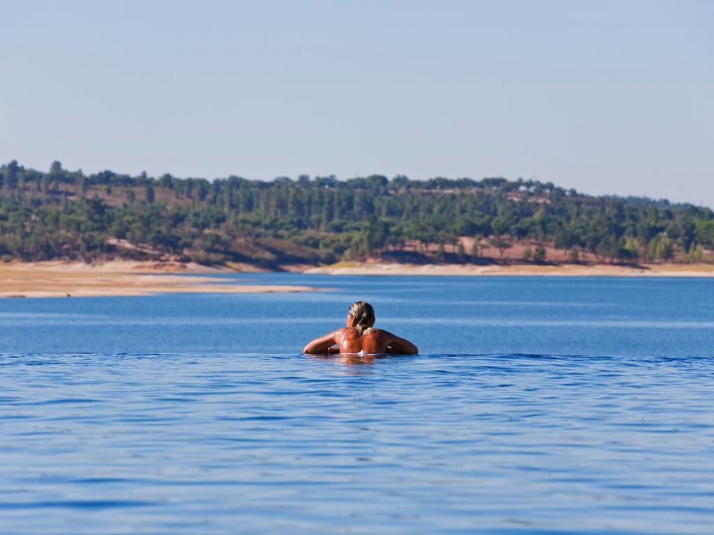 Ap Lago Montargil - Conference & Spa Zewnętrze zdjęcie The photo shows a person swimming in a calm body of water, likely a lake or a river. The swimmer is seen from behind and appears to be enjoying the water on a sunny day. In the background, there's a scenic landscape featuring green trees and a sandy 
