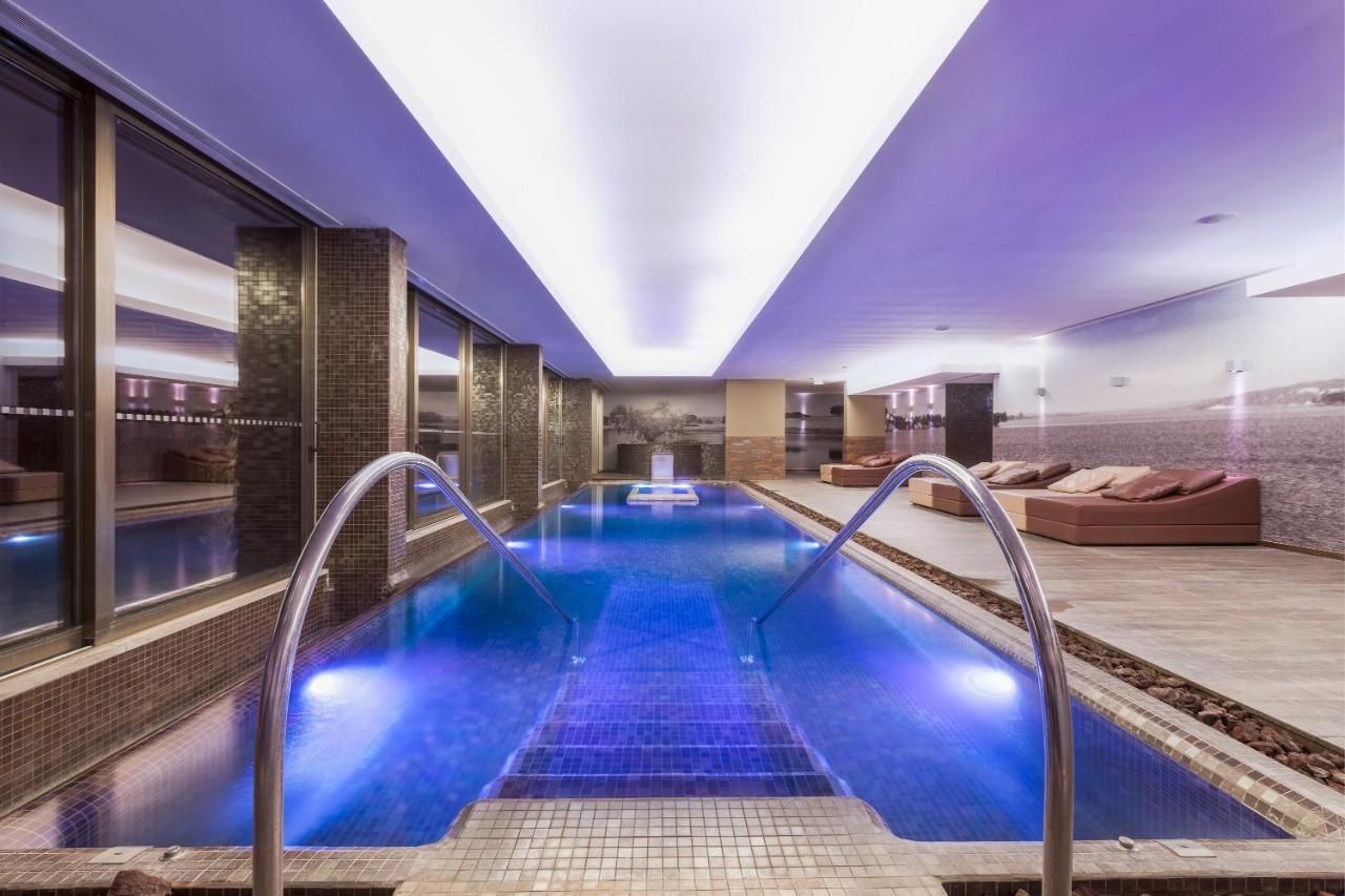 Ap Lago Montargil - Conference & Spa Zewnętrze zdjęcie The photo shows a modern indoor swimming pool area. The pool features a sleek design with steps leading into the water and a gentle curve at the sides. The water is illuminated in a blue hue, enhancing the relaxing atmosphere. Surrounding the pool ar