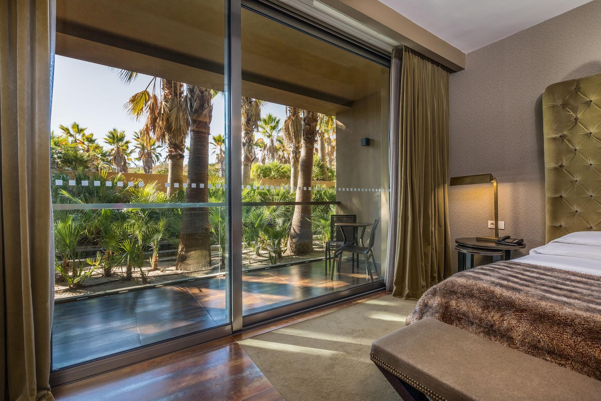 Ap Lago Montargil - Conference & Spa Zewnętrze zdjęcie The photo shows a bedroom with large glass doors that lead out to a balcony or terrace. The view outside features palm trees and lush greenery, indicating a tropical or warm climate. Inside the room, there is a bed, a bedside table with a lamp, and a