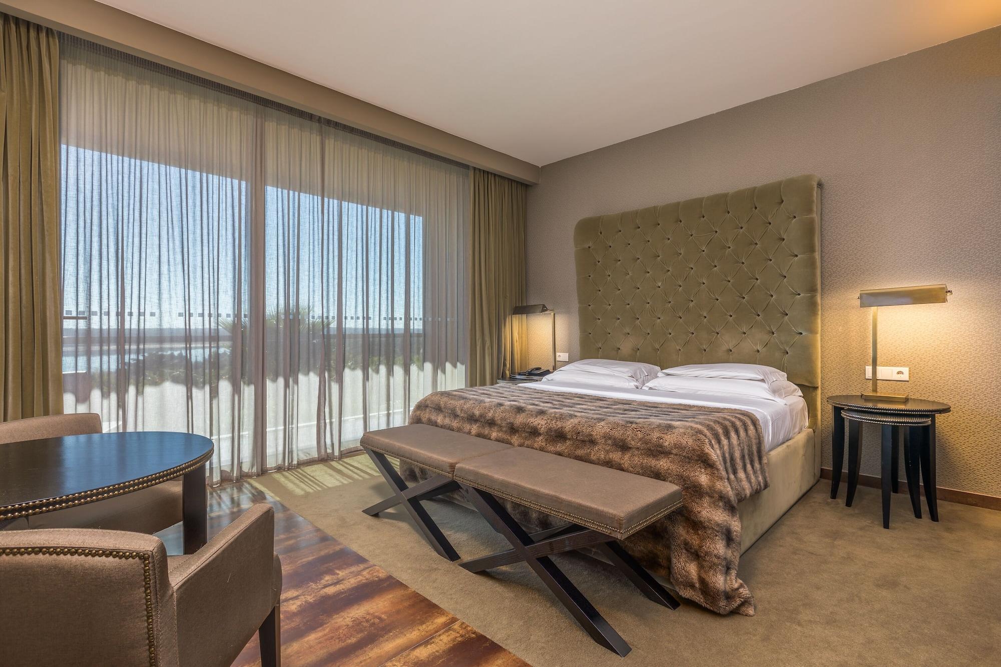 Ap Lago Montargil - Conference & Spa Zewnętrze zdjęcie The photo shows a modern hotel room featuring a large bed with a tufted headboard and a cozy blanket. There are curtains draped over large windows that allow natural light to flow in, providing a view of the outside. A small seating area includes two