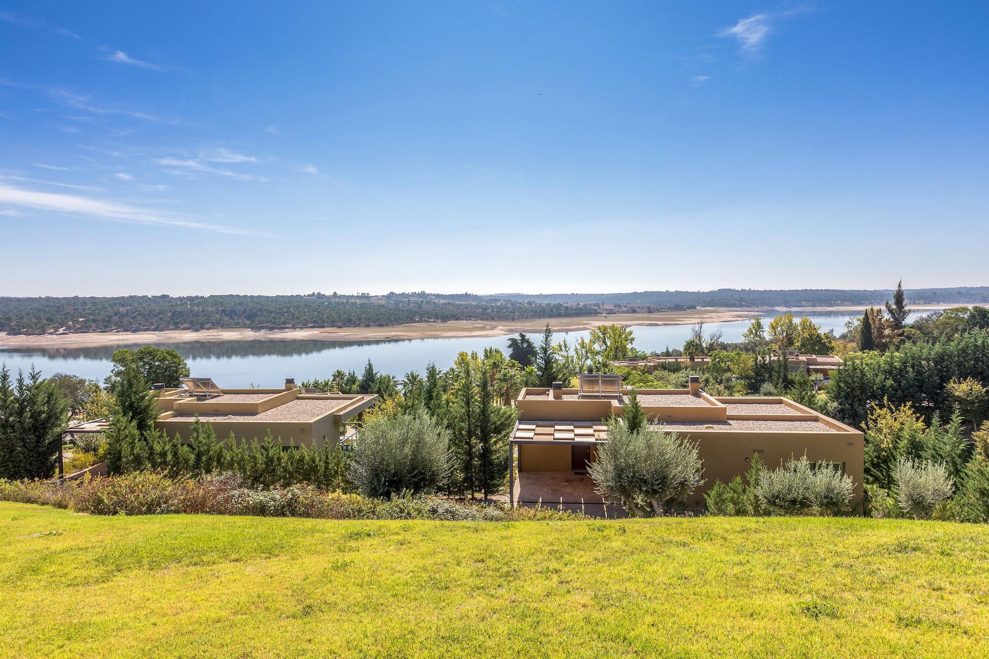Ap Lago Montargil - Conference & Spa Zewnętrze zdjęcie The photo depicts a scenic landscape featuring a calm body of water, possibly a lake or river, surrounded by lush greenery and trees. In the foreground, there are several modern buildings with flat roofs, set against a vibrant blue sky. The overall s
