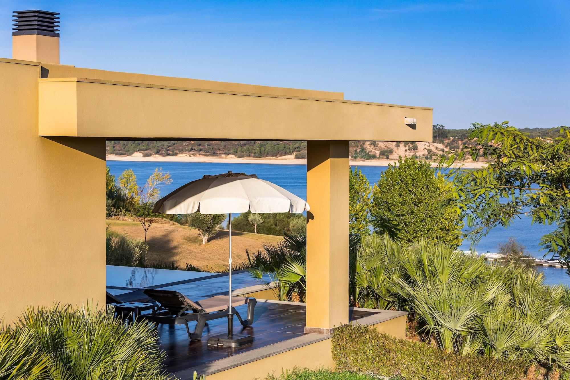 Ap Lago Montargil - Conference & Spa Zewnętrze zdjęcie The photo shows a peaceful outdoor area with a modern architectural structure featuring an overhang. There is a lounge chair positioned beneath a large umbrella, creating a shaded spot to relax. In the background, there is a picturesque view of a bod