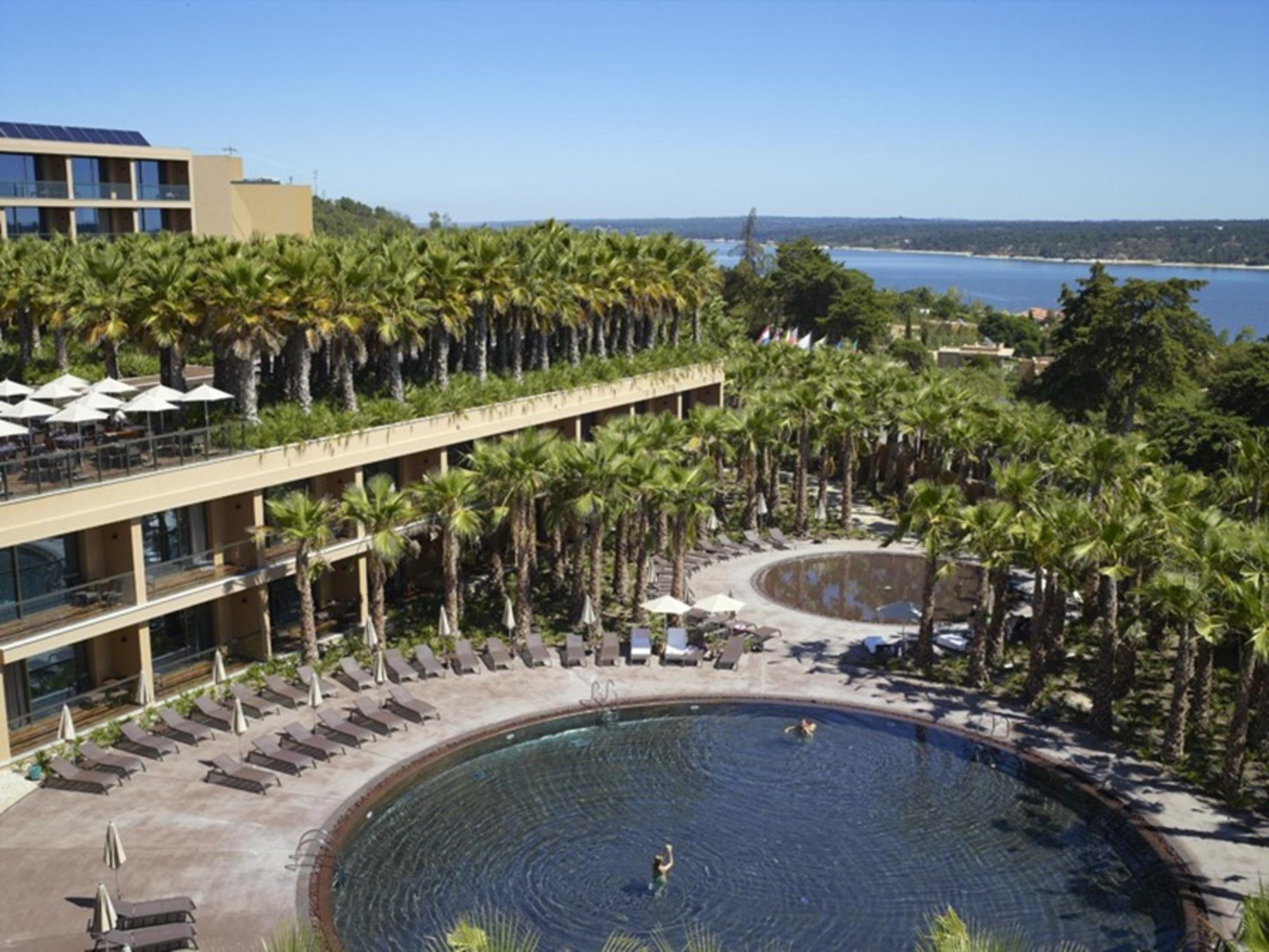Ap Lago Montargil - Conference & Spa Zewnętrze zdjęcie The photo shows a scenic view of a resort area. In the foreground, there are circular pools surrounded by lounge chairs, with a few people enjoying the water. Palm trees line the edges, creating a tropical atmosphere. In the background, there is a mu