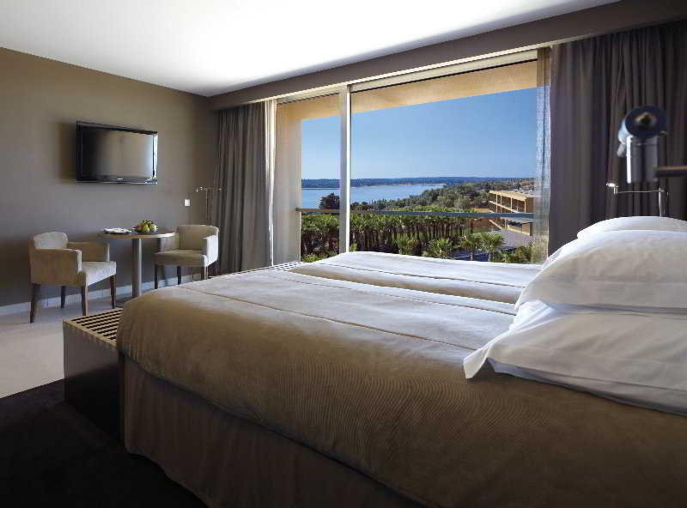 Ap Lago Montargil - Conference & Spa Zewnętrze zdjęcie The photo shows a modern hotel room with a large window offering a view of the sea and mountains. The room features a comfortable bed with white pillows and brown bedding. There is a small sitting area with two chairs and a table, as well as a wall-m