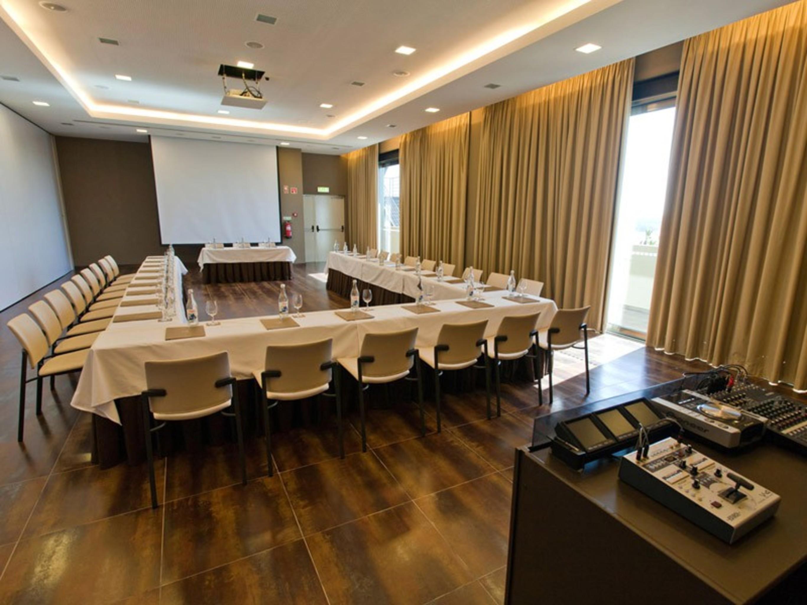 Ap Lago Montargil - Conference & Spa Zewnętrze zdjęcie The photo shows a modern conference room set up for a meeting. There are several tables arranged in a U-shape, surrounded by chairs. A large screen and projector are positioned at the front, with a table for speakers or presenters. The room has large