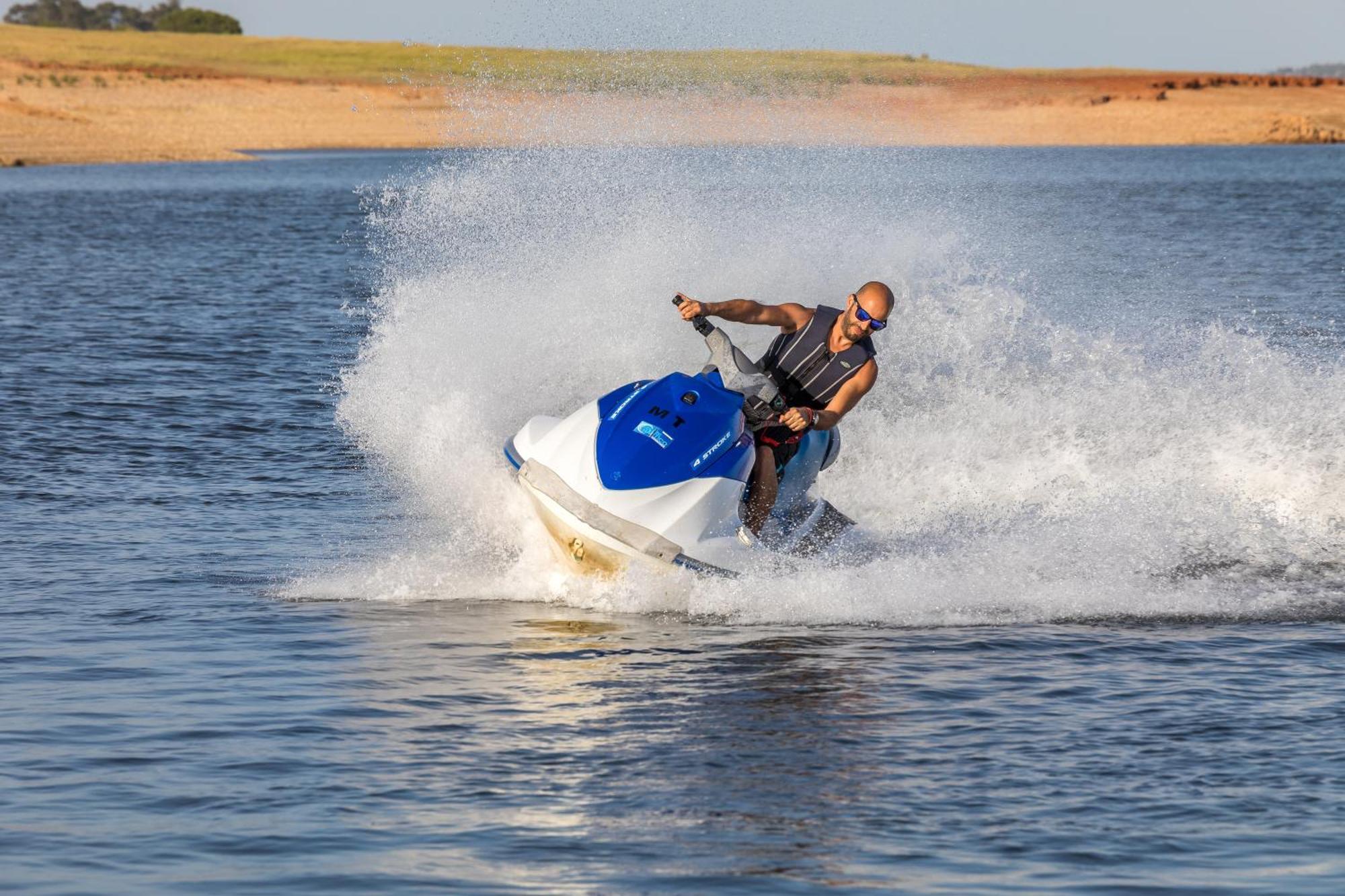 Ap Lago Montargil - Conference & Spa Zewnętrze zdjęcie The photo shows a person riding a jet ski over a body of water. The rider is wearing sunglasses and a life vest, leaning forward as the jet ski accelerates, creating a spray of water around them. The background features a natural landscape, likely a 