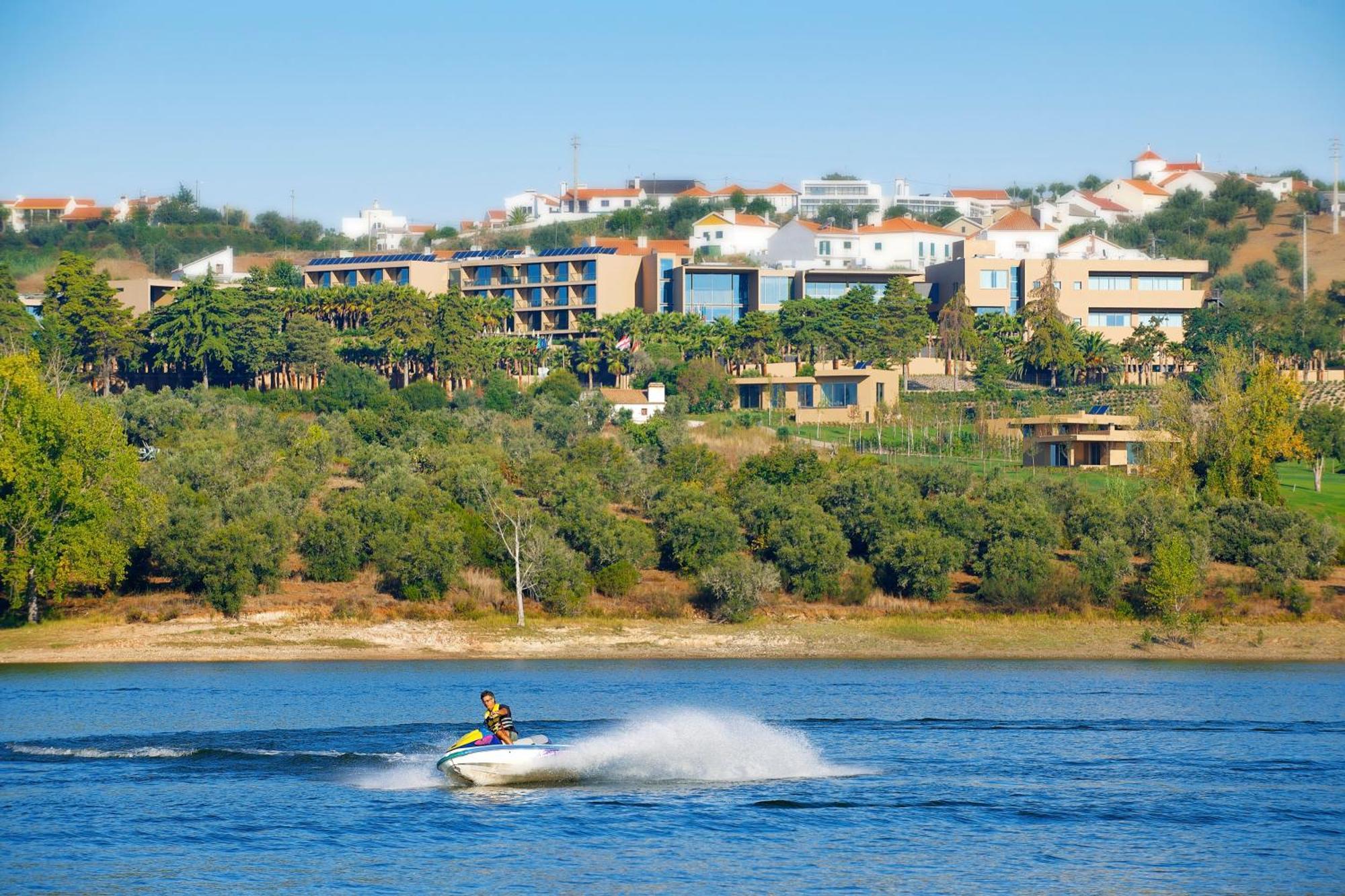 Ap Lago Montargil - Conference & Spa Zewnętrze zdjęcie The photo shows a person riding a jet ski on a body of water, creating a spray as they speed across the surface. In the background, there are modern buildings and a hillside with trees and greenery. The sky is clear and blue, suggesting a bright and 