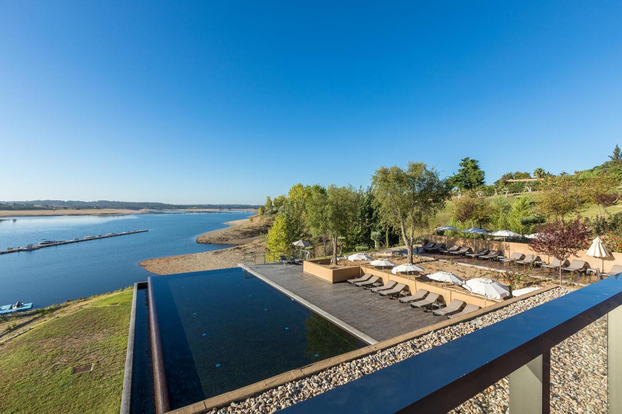 Ap Lago Montargil - Conference & Spa Zewnętrze zdjęcie The photograph depicts a scenic view of a tranquil water body, likely a lake or river, bordered by lush greenery. In the foreground, there is an infinity pool that blends seamlessly with the water beyond. Comfortable lounge chairs are arranged on a w